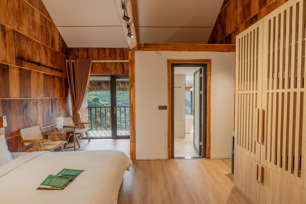 Una habitación moderna con suelo de madera, una cama con un libro verde, un armario de madera y un balcón con vistas.