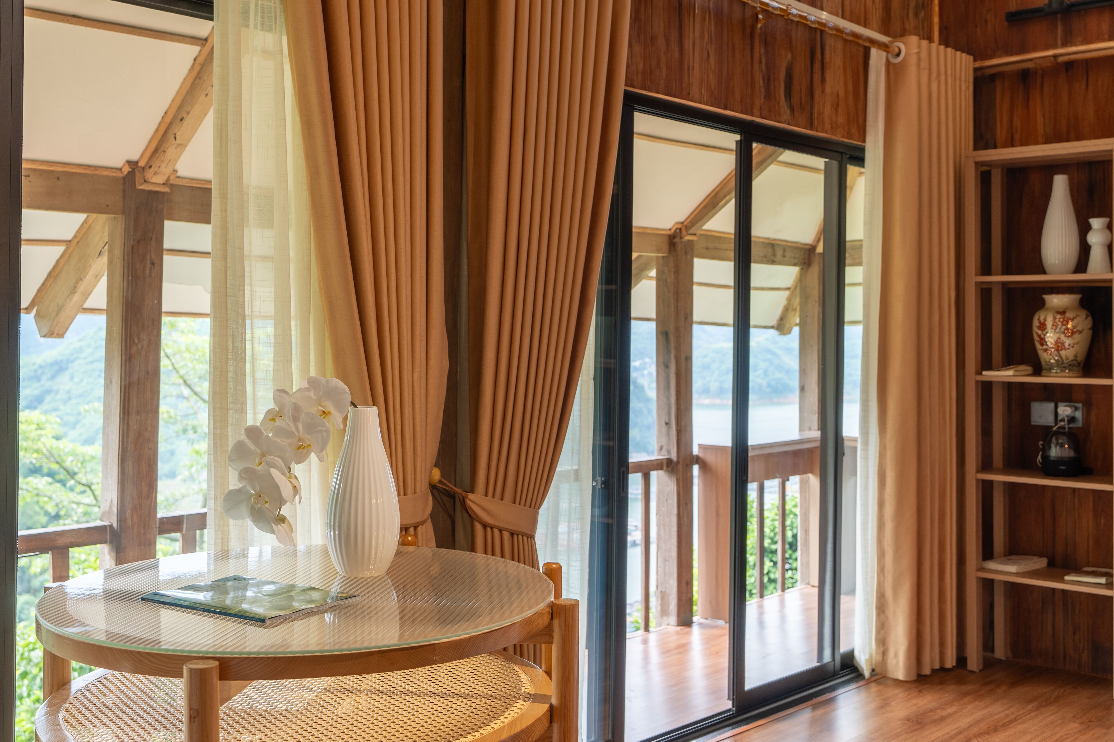 A room with a glass table, wicker chair, white vase, and a view of the outdoors.