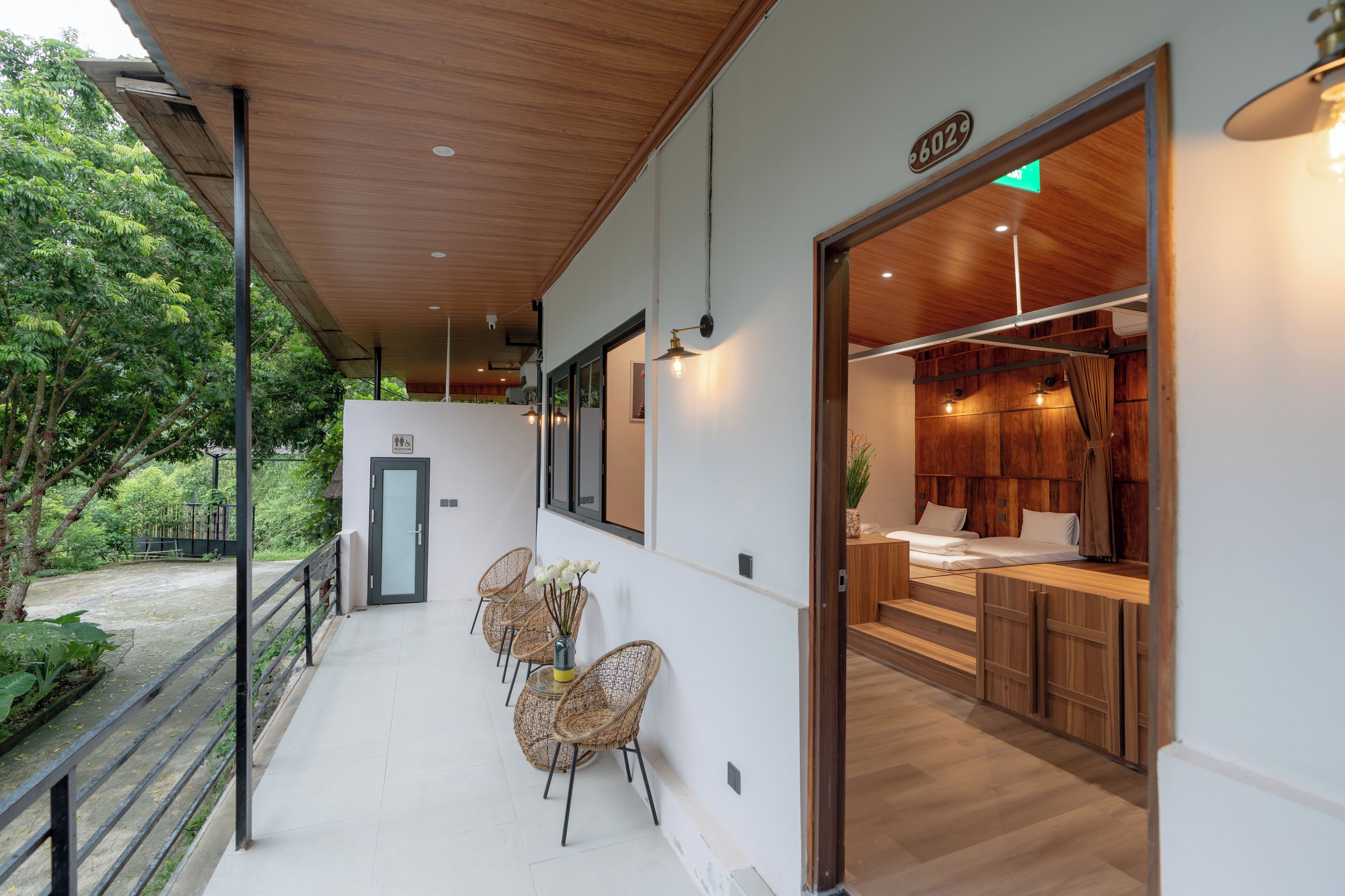 A modern hotel room with a wooden ceiling, a balcony with wicker chairs and a table, and a view of the greenery outside.