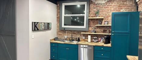 A modern kitchen with teal cabinets, a stainless steel dishwasher, and a brick wall.