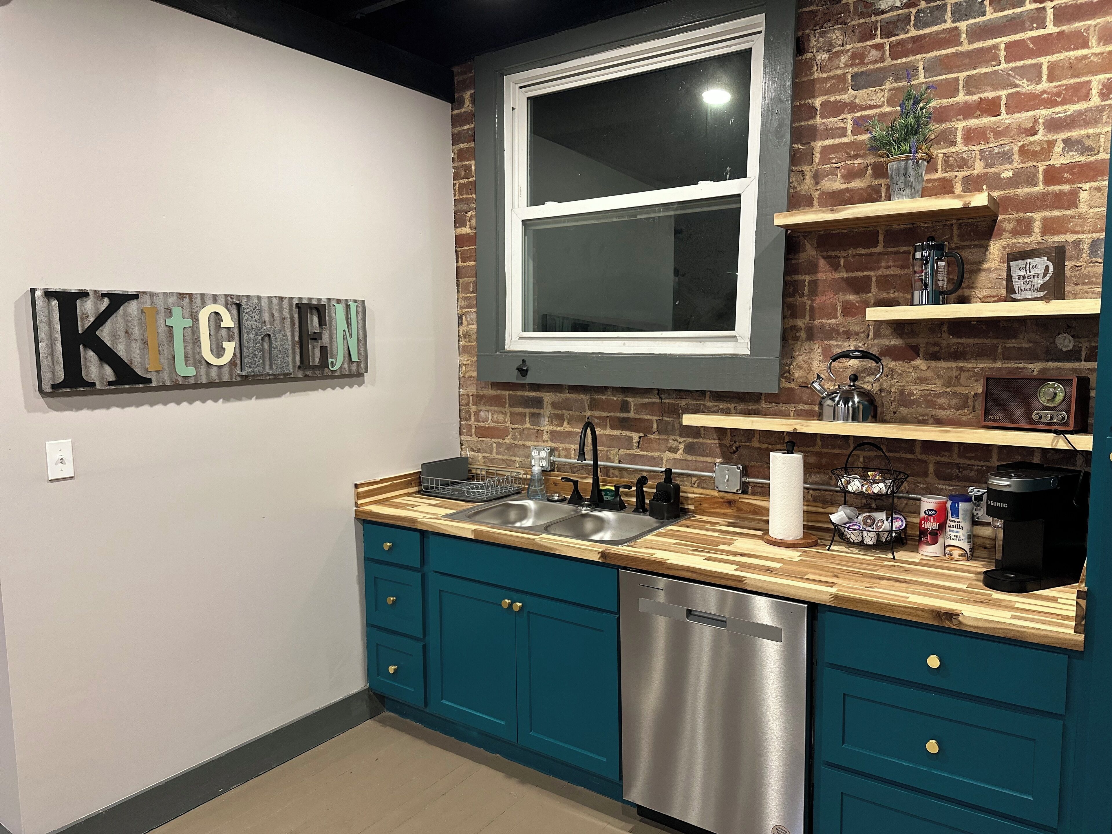 A modern kitchen with teal cabinets, a stainless steel dishwasher, a wooden countertop, and a brick wall.