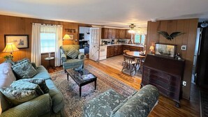 A cozy living room with a sofa, coffee table, and a dining area with chairs. A kitchen is visible in the background. - Orleans Beach Retreat (Orleans)