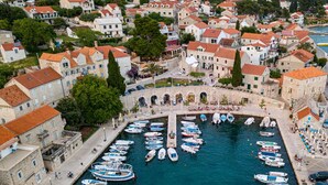 Une ville côtière aux bâtiments en pierre, un port de plaisance où sont amarrés de nombreux bateaux, et une place centrale animée, avec des passants et des terrasses.