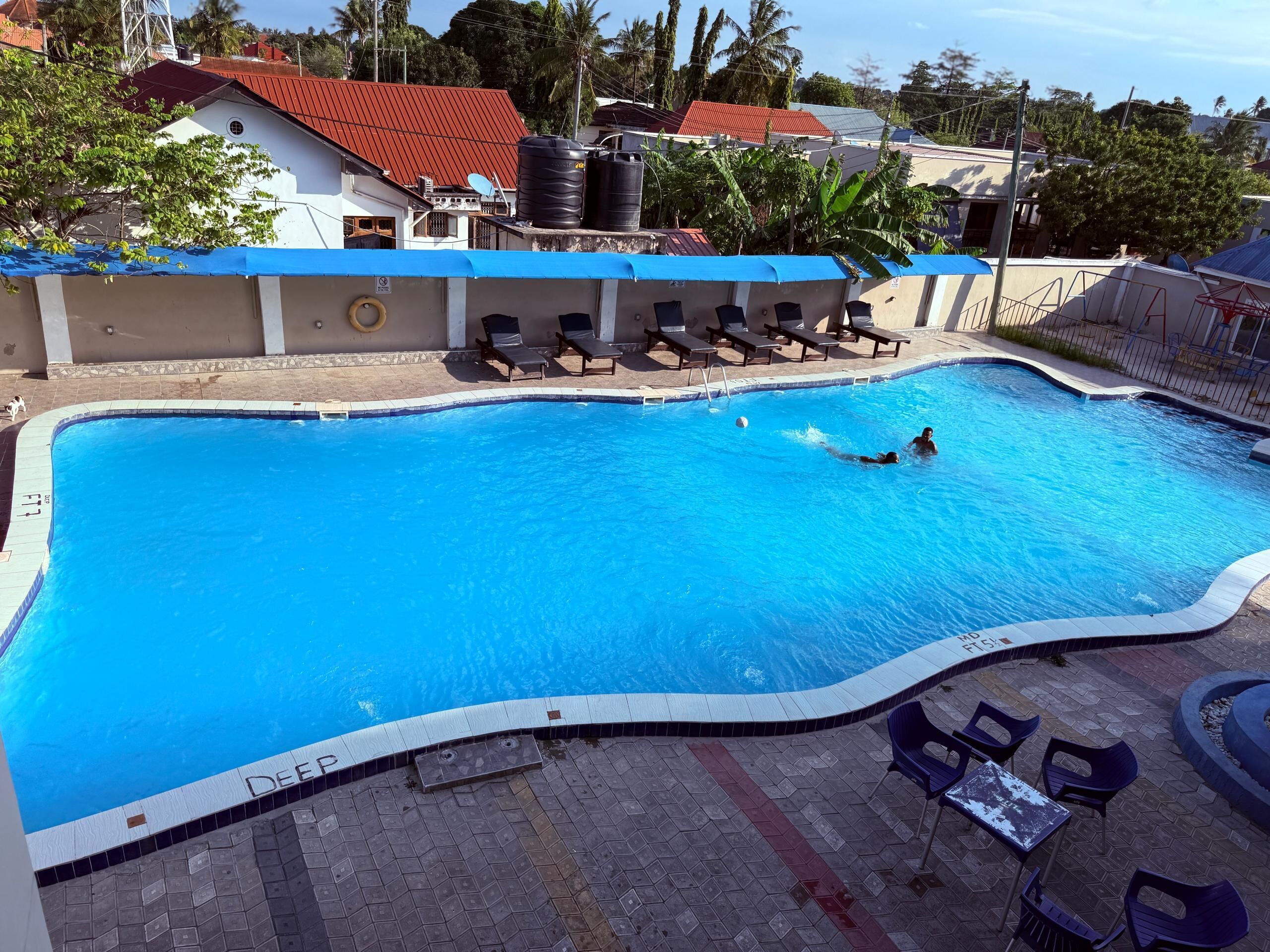 Ein Schwimmbecken mit gefliestem Sonnendeck, Liegestühlen und einem umgebenden Gebäude mit rotem Dach.