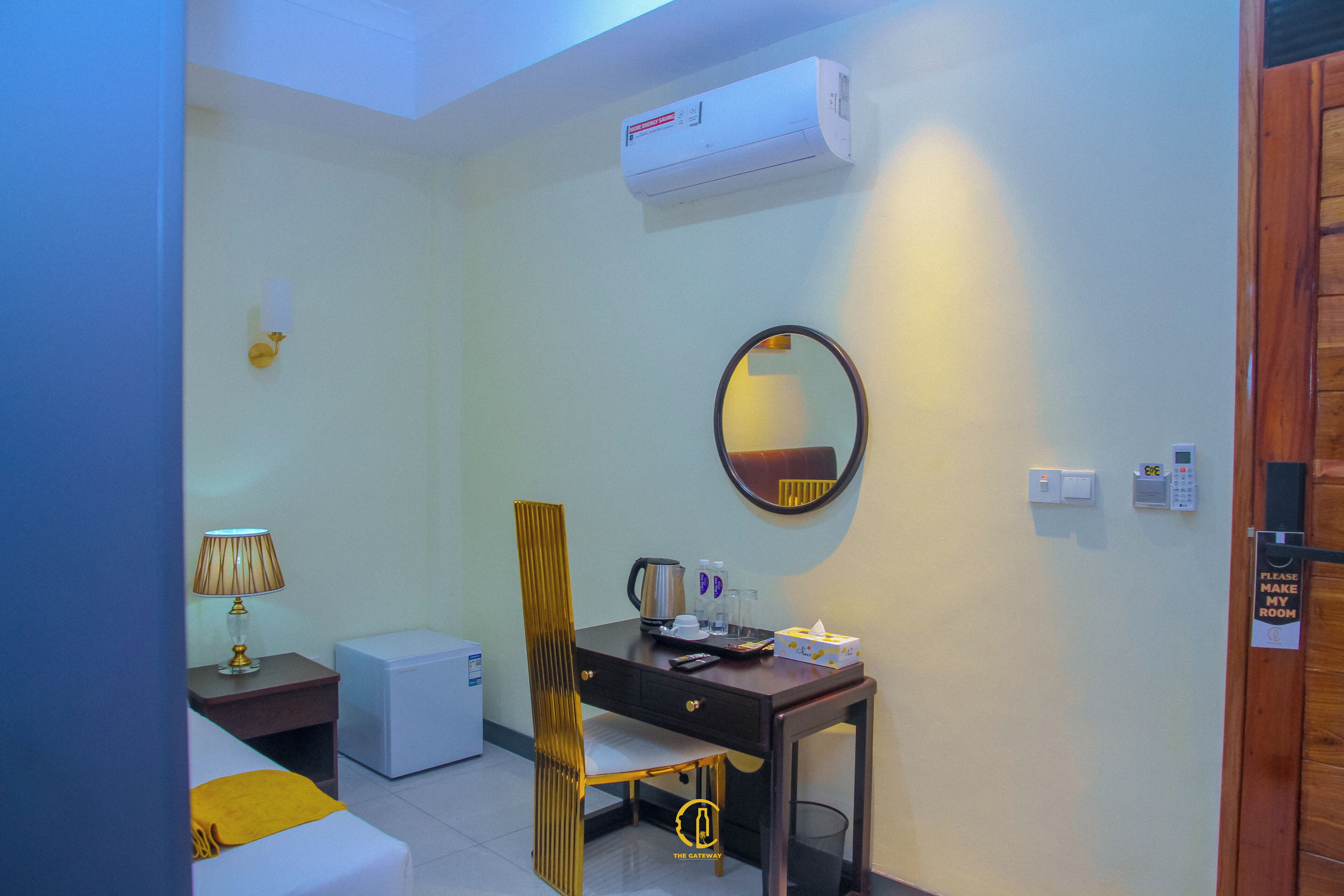 Een compacte hotelkamer met een bureau, stoel en een klein tafeltje met een lamp. Een airconditioner is aan de muur bevestigd en een spiegel hangt boven het bureau.