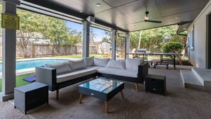 Un espace extérieur aménagé avec une zone salon couverte, donnant sur la piscine, comprenant un canapé blanc et une table basse en verre.