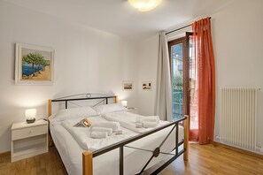 A neatly made bed with white linens and towels, a bedside table with a lamp, a framed painting on the wall, and a window with orange curtains.