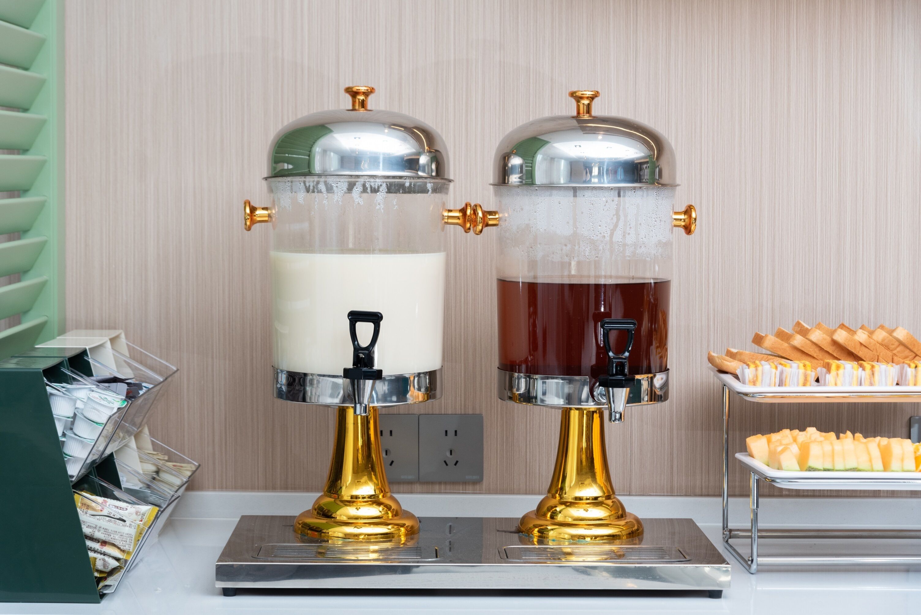 Twee automaten voor zelfbediening met gouden voetstukken en glazen containers, een schaal met gebak en een stapel kranten.