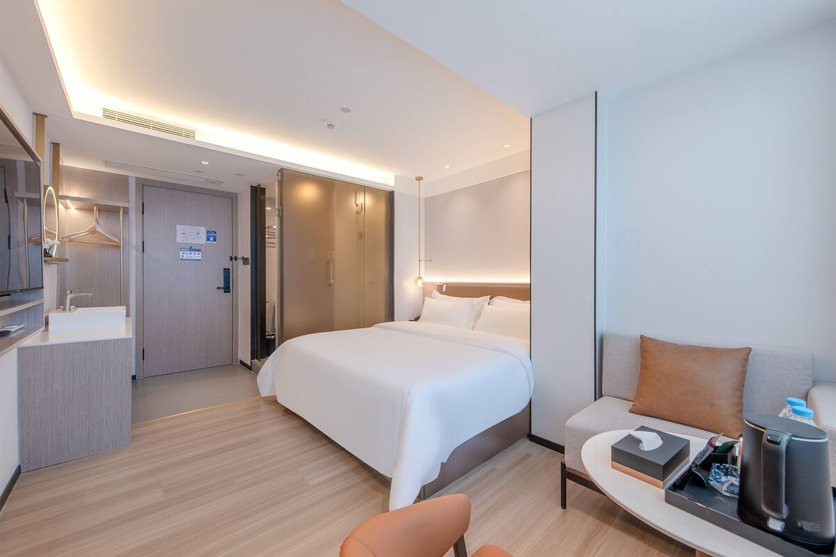 A modern hotel room with a large bed, a sofa, a desk with a coffee maker, and a bathroom with a sink and mirror.