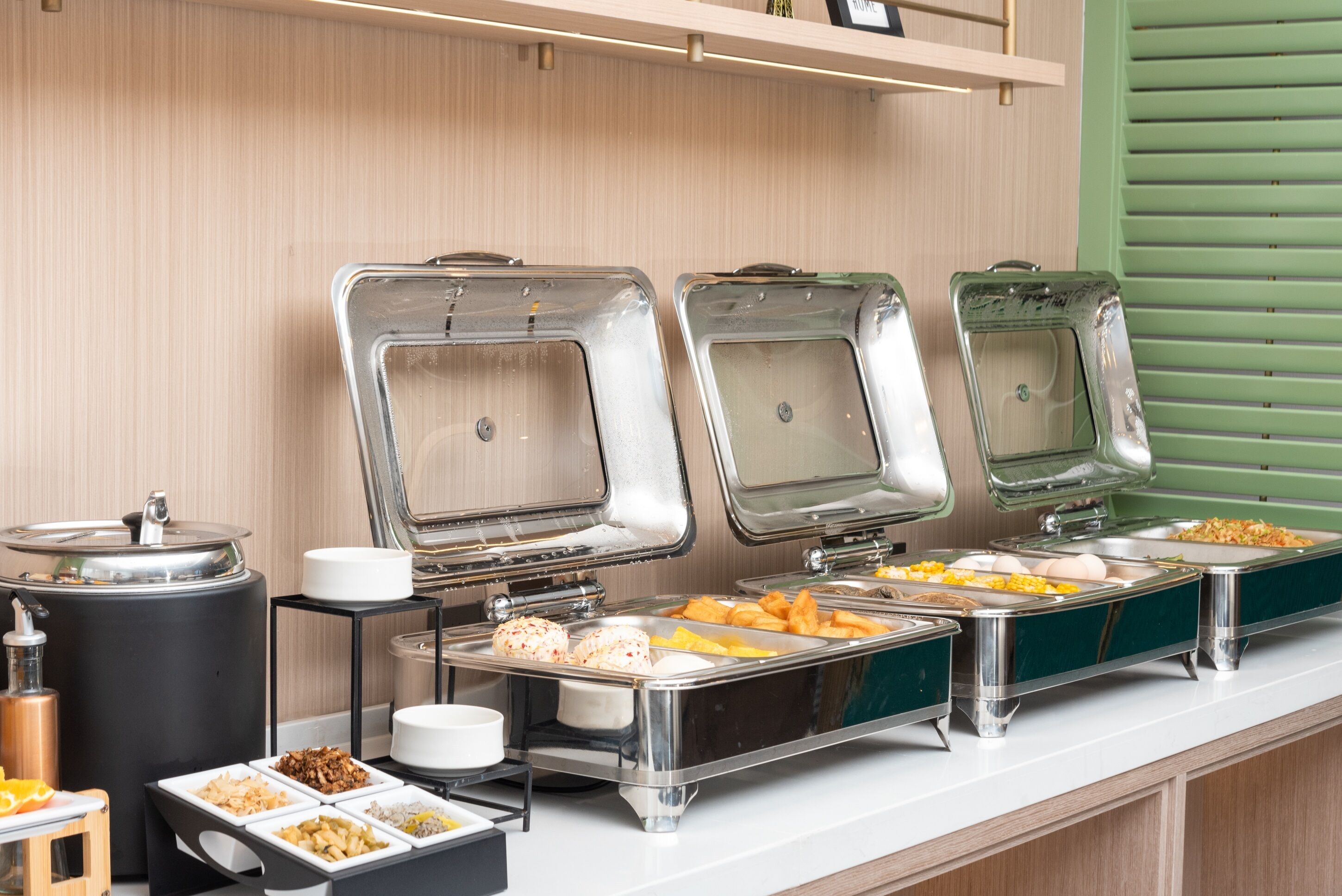 A buffet setup with various food items in metal containers, a coffee station, and a small table with a cutting board and knife.