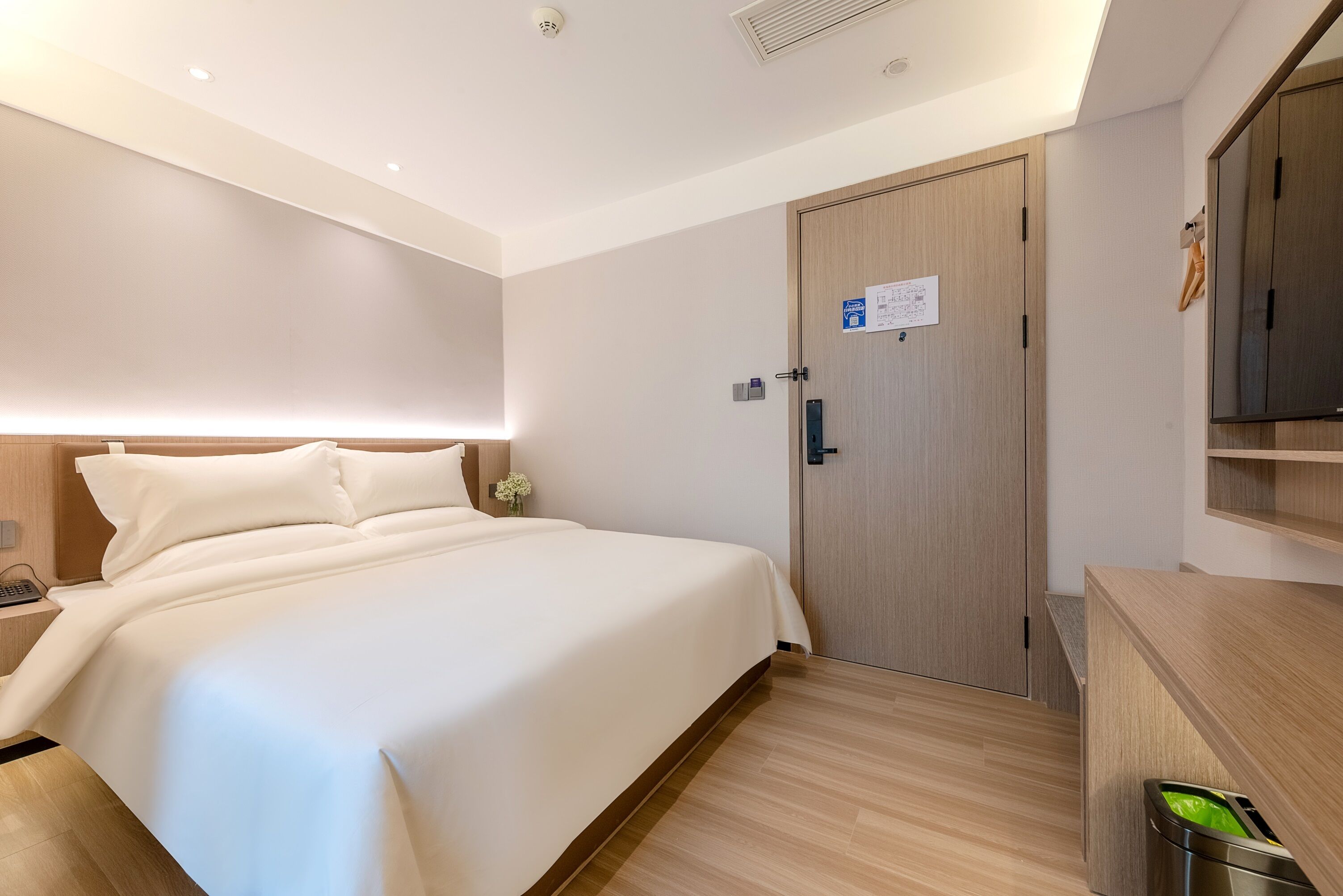 Una habitación de hotel moderna con una cama grande, un armario de madera, un escritorio pequeño y un cesto de basura.
