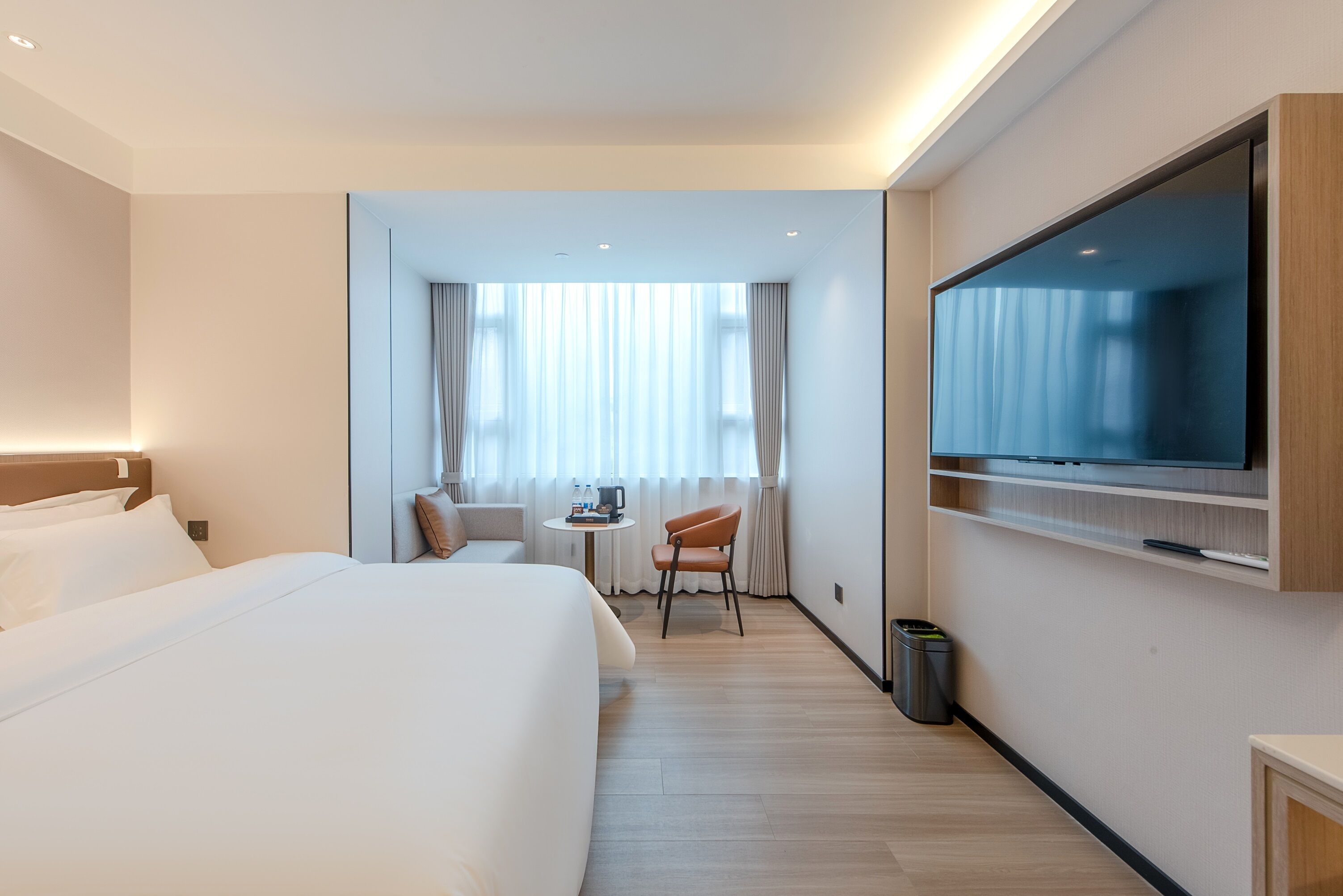 A hotel room with a large bed, a TV mounted on the wall, a small table with a chair, and a window with curtains.