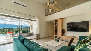 Un salon moderne avec un grand canapé vert, une table basse blanche et un téléviseur fixé au mur. Il y a un balcon avec vue sur les montagnes et une piscine.