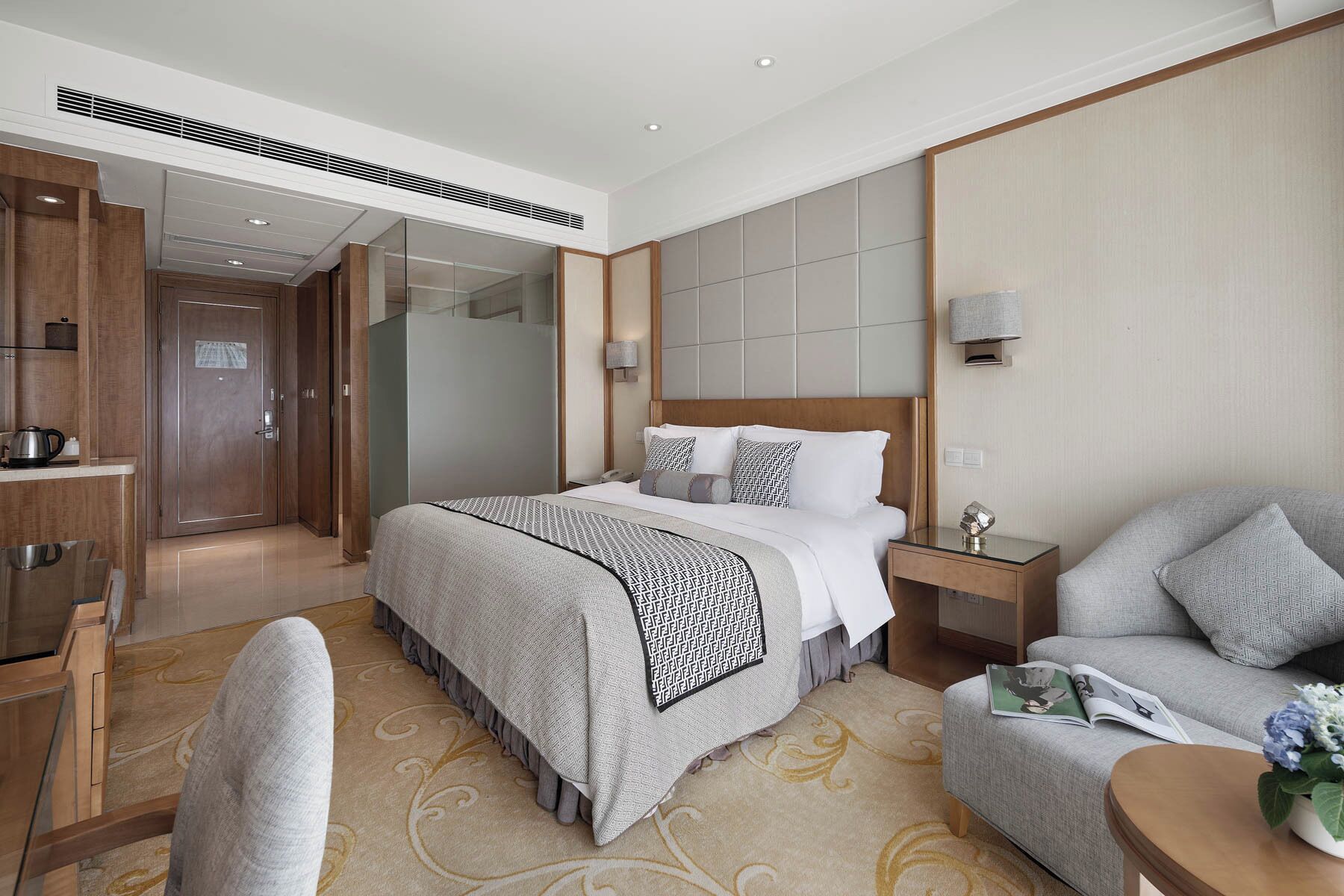 A modern hotel room with a large bed, a sitting area with a sofa and armchair, a desk with a lamp, and a walk-in closet.