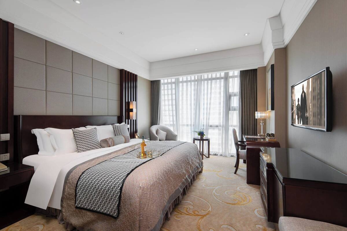 A hotel room with a large bed, a desk, a chair, and a view of the city through large windows.
