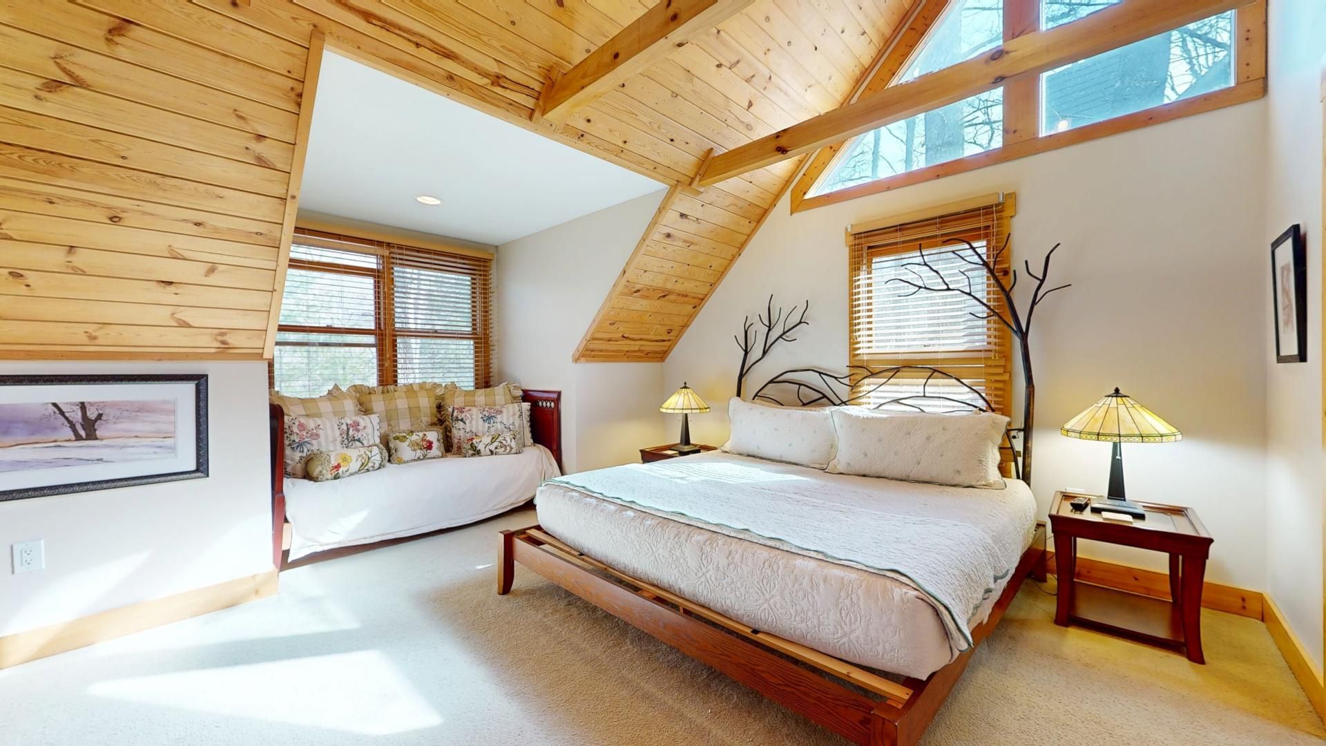 A bedroom with a sloped ceiling, a bed, two bedside tables with lamps, a window with blinds, and a framed picture on the wall.