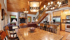Un vaste salon doté d’une grande table à manger en bois, de sièges confortables, d’une cheminée et d’un escalier menant à un niveau supérieur.