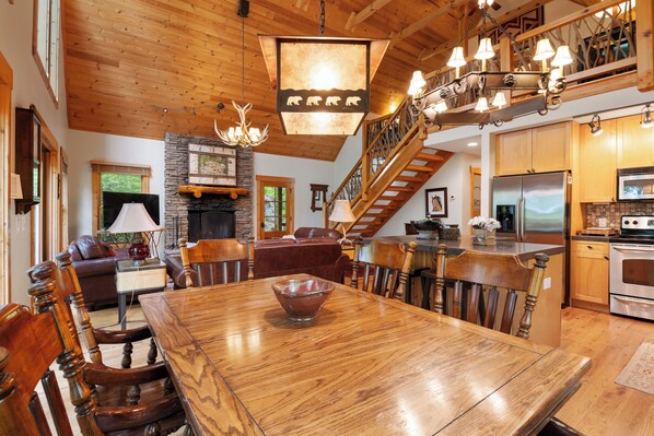 Amplia sala de estar con una gran mesa de madera para comer, asientos cómodos, una chimenea y una escalera que sube a un nivel superior.
