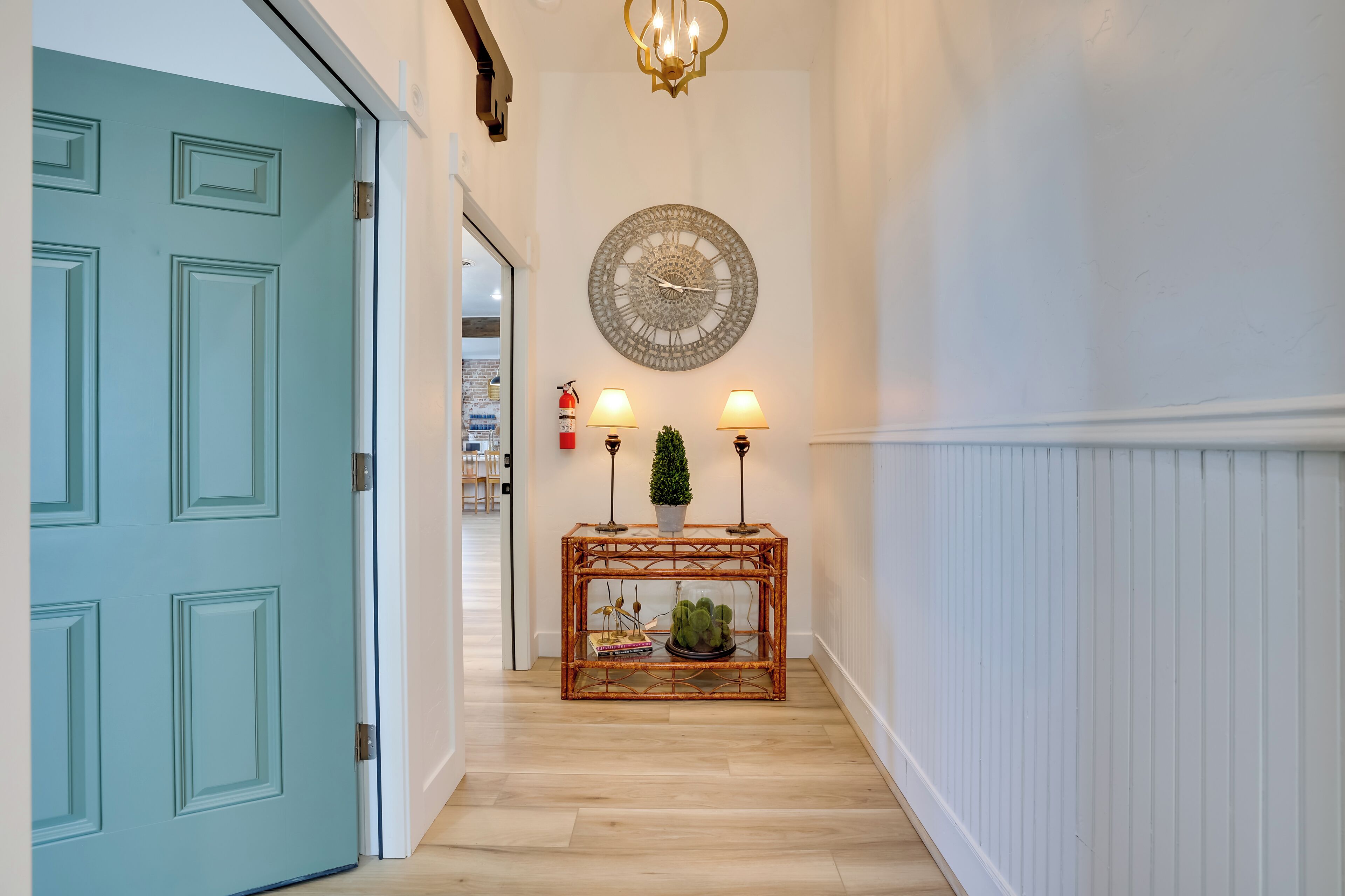 Un pasillo con una puerta color turquesa, un reloj decorativo en la pared, una consola con lámparas y una planta pequeña.