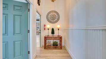 Un pasillo con una puerta color turquesa, un reloj decorativo en la pared, una consola con lámparas y una planta pequeña.
