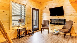 Ein gemütliches Holzhaus-Interieur mit Kamin, zwei Sesseln, einem kleinen Tisch mit Lampe und einem an der Wand befestigten Fernseher.