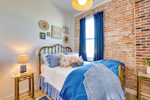 Een slaapkamer met een bakstenen muur, een bed met blauw beddengoed, een messing bedframe, een nachtkastje met een lamp en lijstjes op de muur.