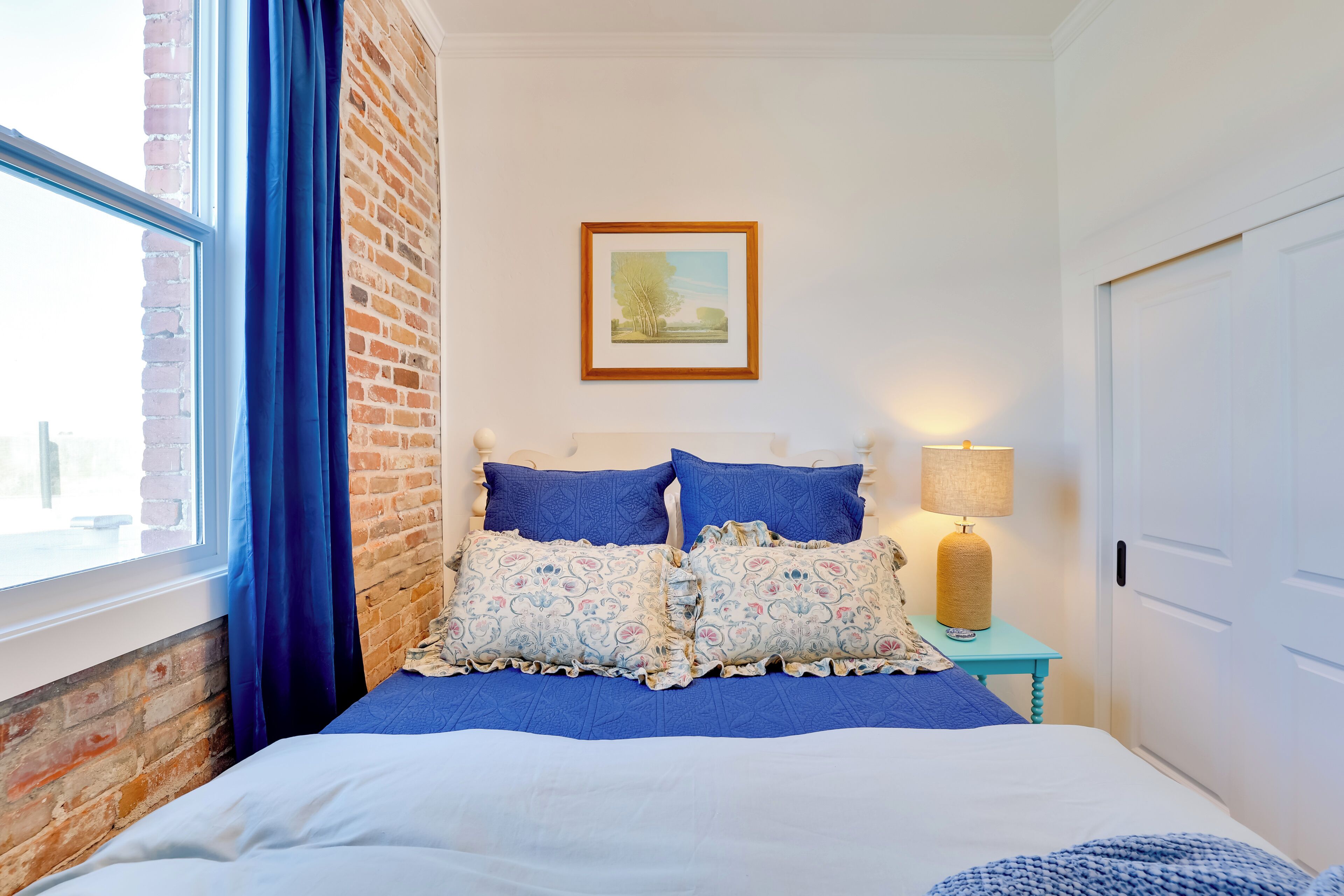 Un dormitorio con pared de ladrillo, un ventanal con cortinas azules, una cama con ropa de cama azul, una mesita de noche con una lámpara y un cuadro enmarcado en la pared.