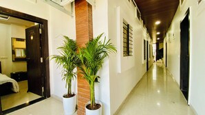 Un pasillo con piso pulido, plantas en macetas, un pilar de madera y una puerta que da a una habitación con cama y cómoda.