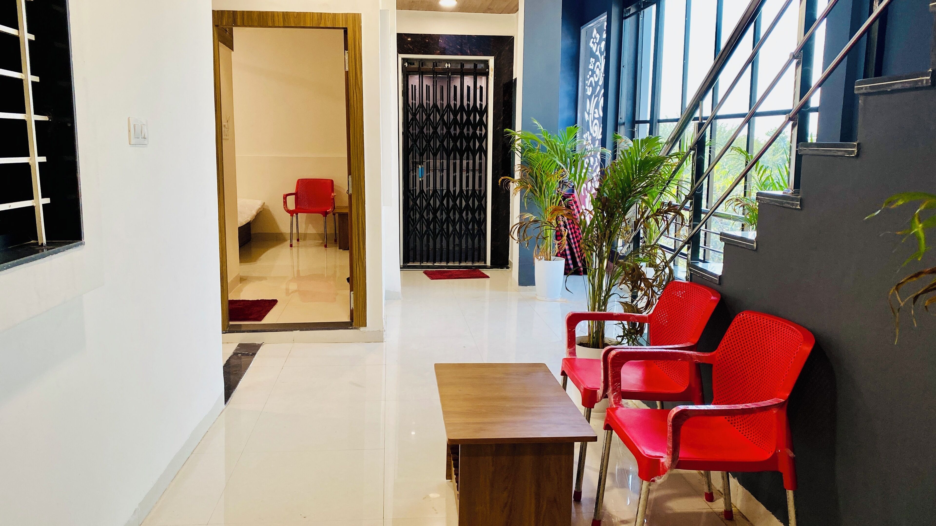 A modern hotel lobby with a wooden reception desk, red chairs, a large potted plant, and a view of the outdoors through large windows.