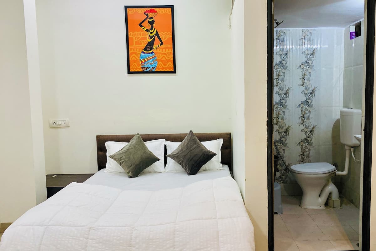 Ein Schlafzimmer mit Bett, Kissen, einem Nachttisch, einem Wandbild und einem Badezimmer mit Toilette und Duschvorhang.