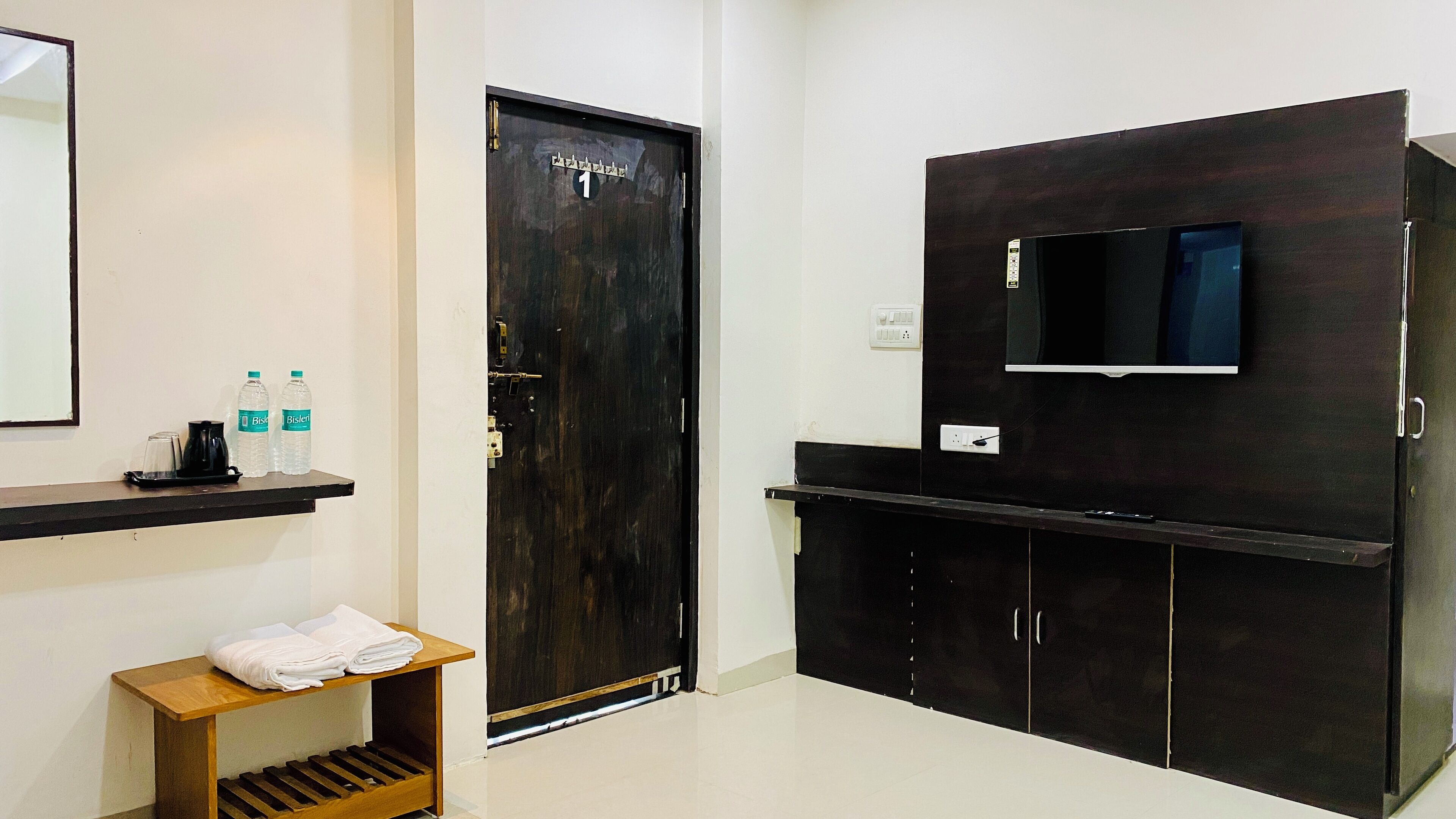 Quarto de hotel com guarda-roupa de madeira escura, televisão de ecrã plano na parede, uma pequena mesa com toalhas e uma estante com garrafas de água e máquina de café.