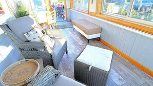 Une pièce baignée de lumière naturelle, meublée de mobilier en osier, dotée de grandes fenêtres et offrant une vue sur l’extérieur.