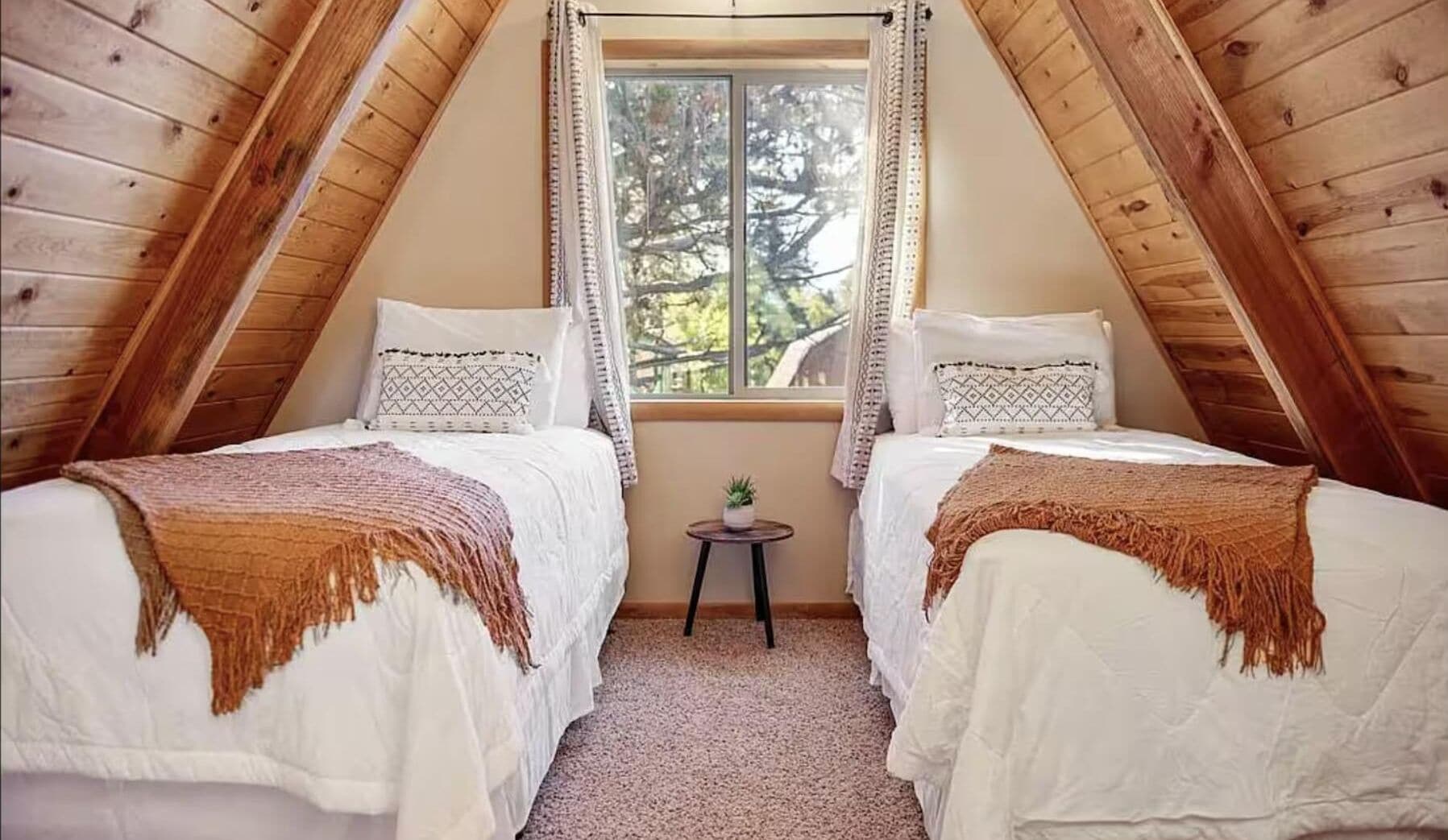 Une chambre mansardée chaleureuse, avec deux lits, une petite table et une fenêtre donnant sur les arbres.