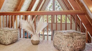 A room with a sloped ceiling, wooden beams, a window with a view of trees, a white vase with pampas grass, and two patterned ottomans. - Charming A-Frame home,  wood fireplace,  7 miles to The Village and ski resorts. (Sugarloaf)
