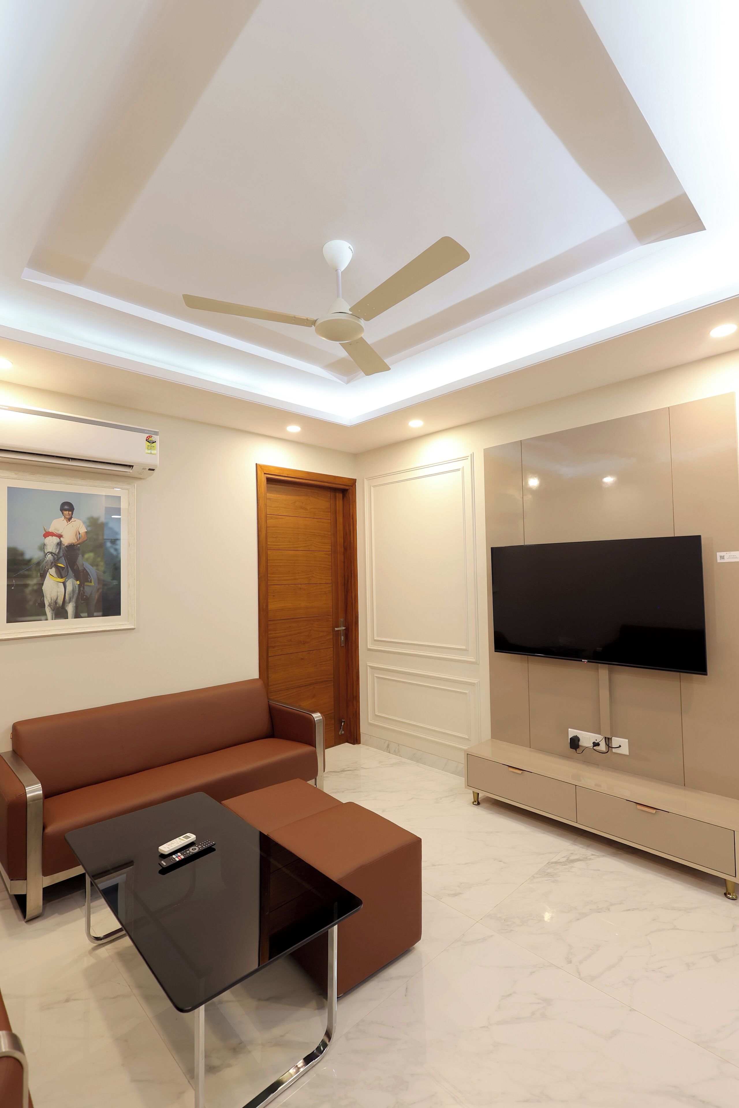 Un salon moderne avec un canapé marron, une table basse en verre, un ventilateur de plafond, un téléviseur fixé au mur et une porte en bois.
