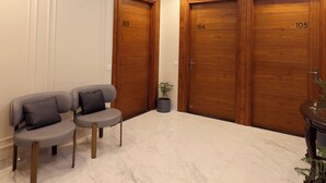 Un hall de hotel con un abanico de techo, puertas de madera con los números 103, 104 y 105, un rincón de descanso con dos sillones y una planta en maceta.