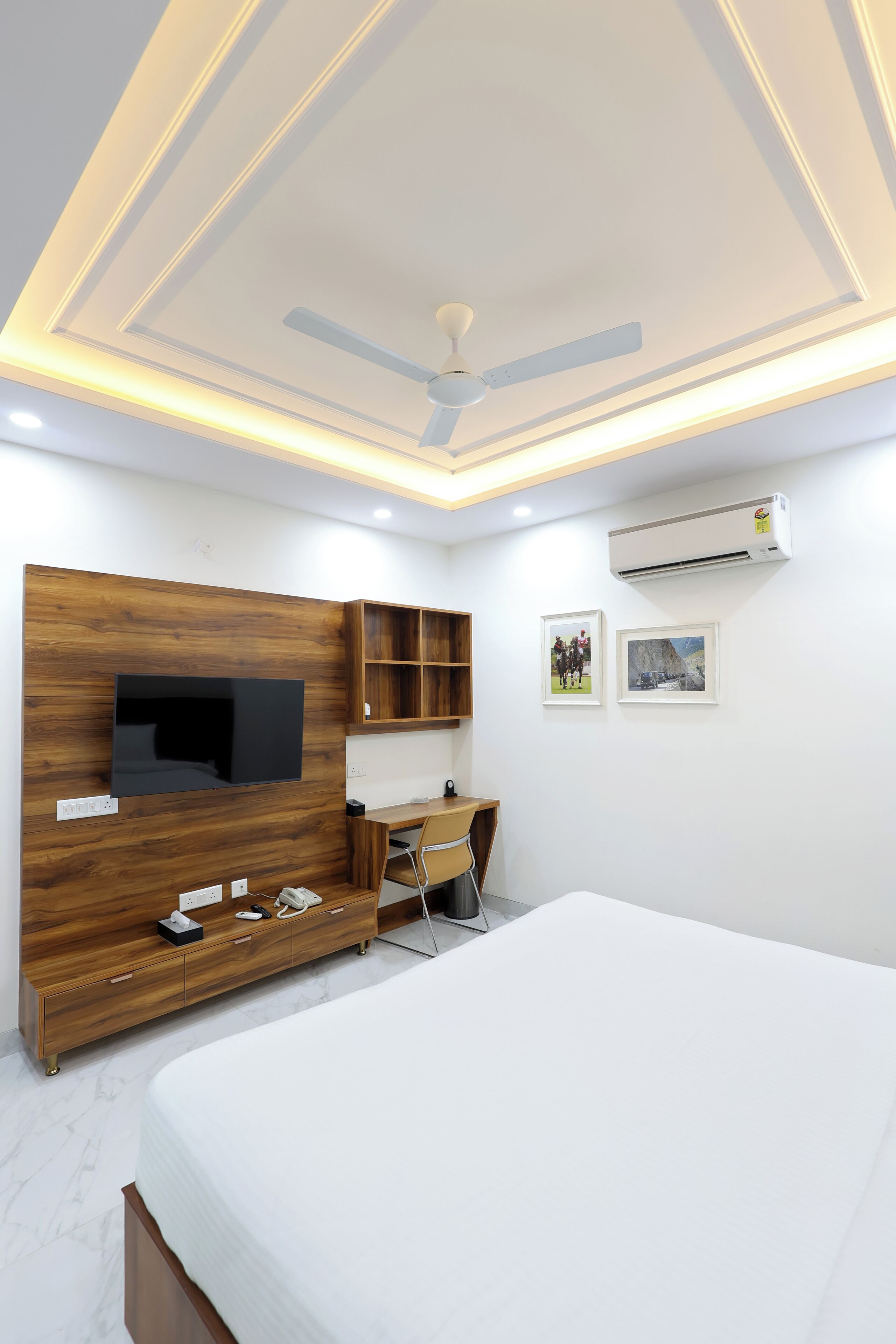 A bedroom with a bed, a TV mounted on a wooden wall, a desk with a chair, and an air conditioning unit on the wall.
