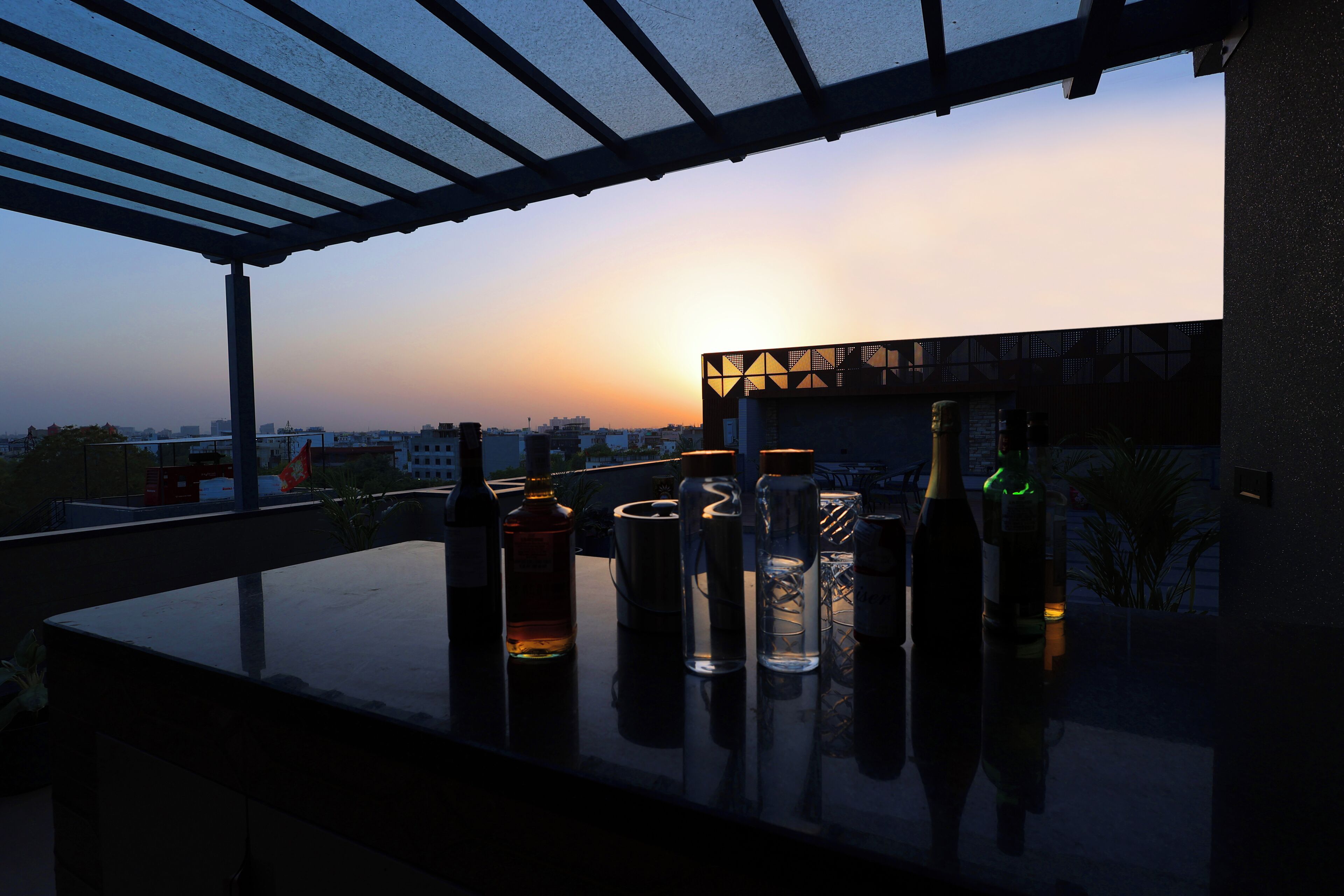 Een dakterras met flessen en glazen, een stadszicht op de achtergrond en een zonsondergang.