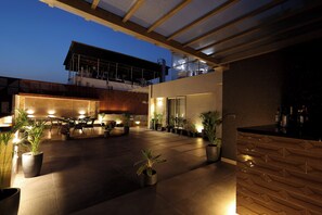 A modern outdoor dining area with potted plants, a covered seating area, and a view of a building with a balcony. - Escape - Stays and Serviced Apartments (Gurugram)