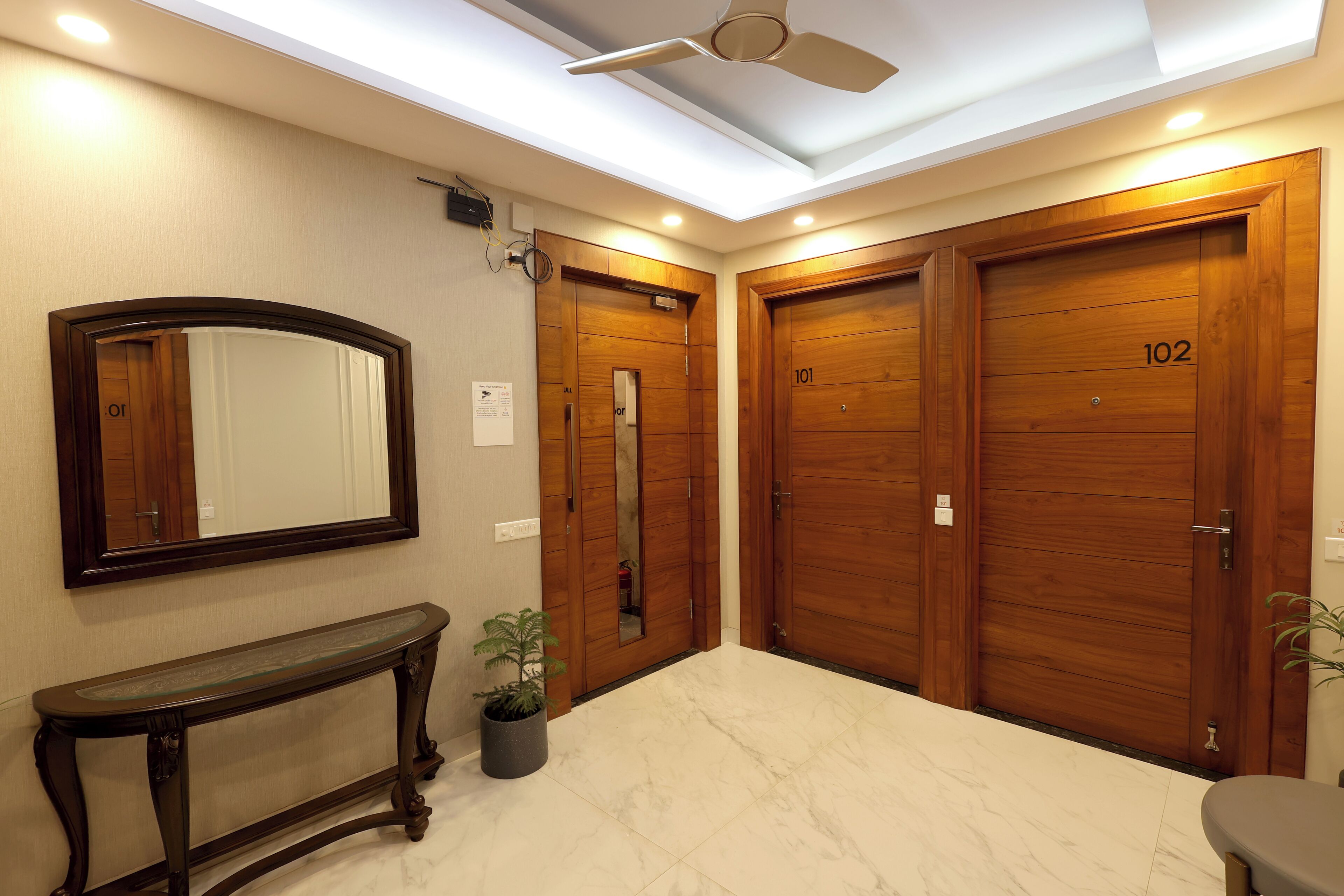 Un hall d’hôtel avec une double porte en bois portant les numéros 101 et 102, un sol en marbre, une console, une plante en pot et un ventilateur de plafond.