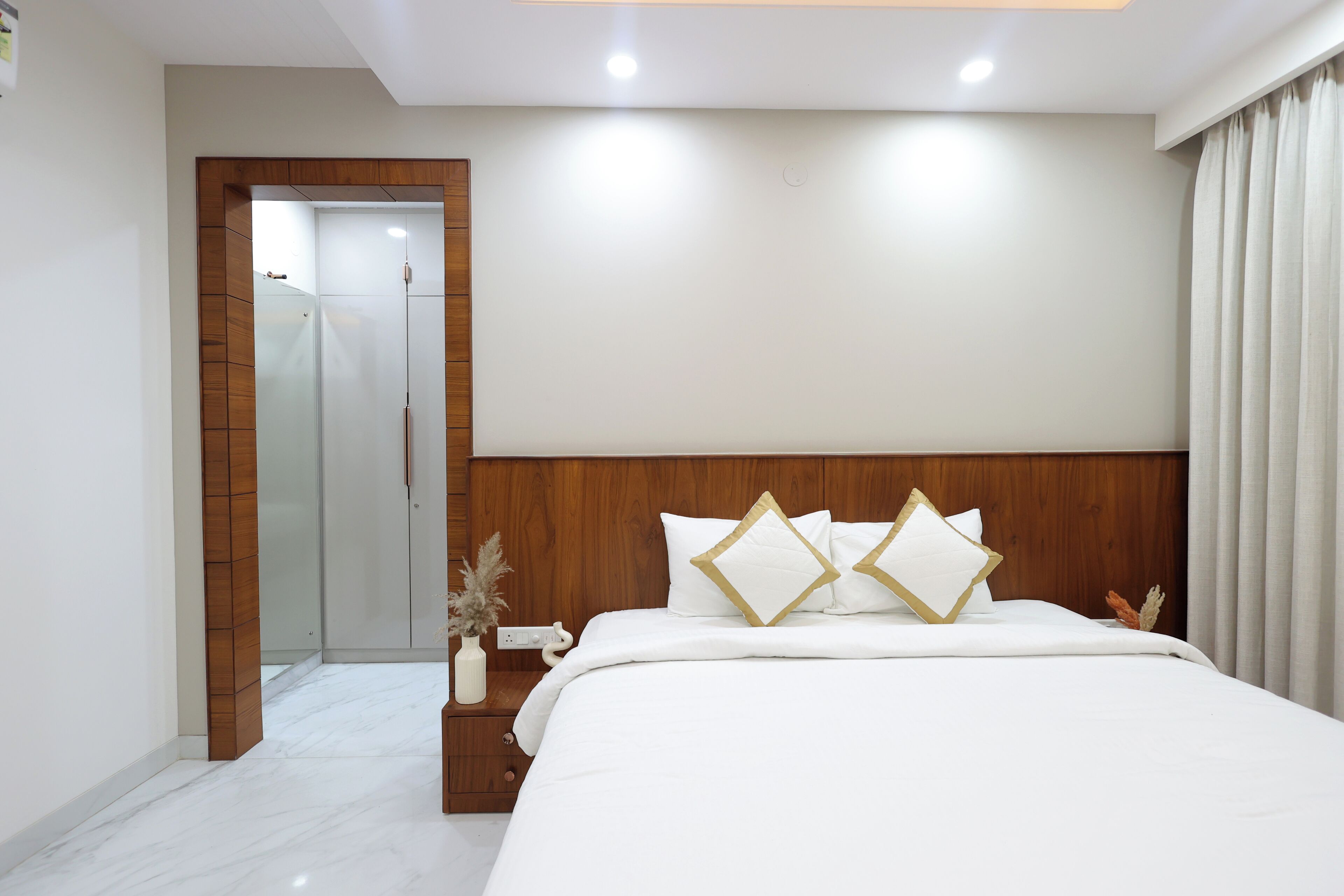Habitación de hotel con cabecera de madera, cama grande con ropa de cama blanca y detalles dorados, mesita de noche con un florero y puerta que da a un baño.