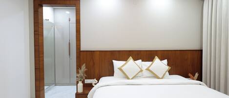 Habitación de hotel con cabecera de madera, cama grande con ropa de cama blanca y detalles dorados, mesita de noche con un florero y puerta que da a un baño.