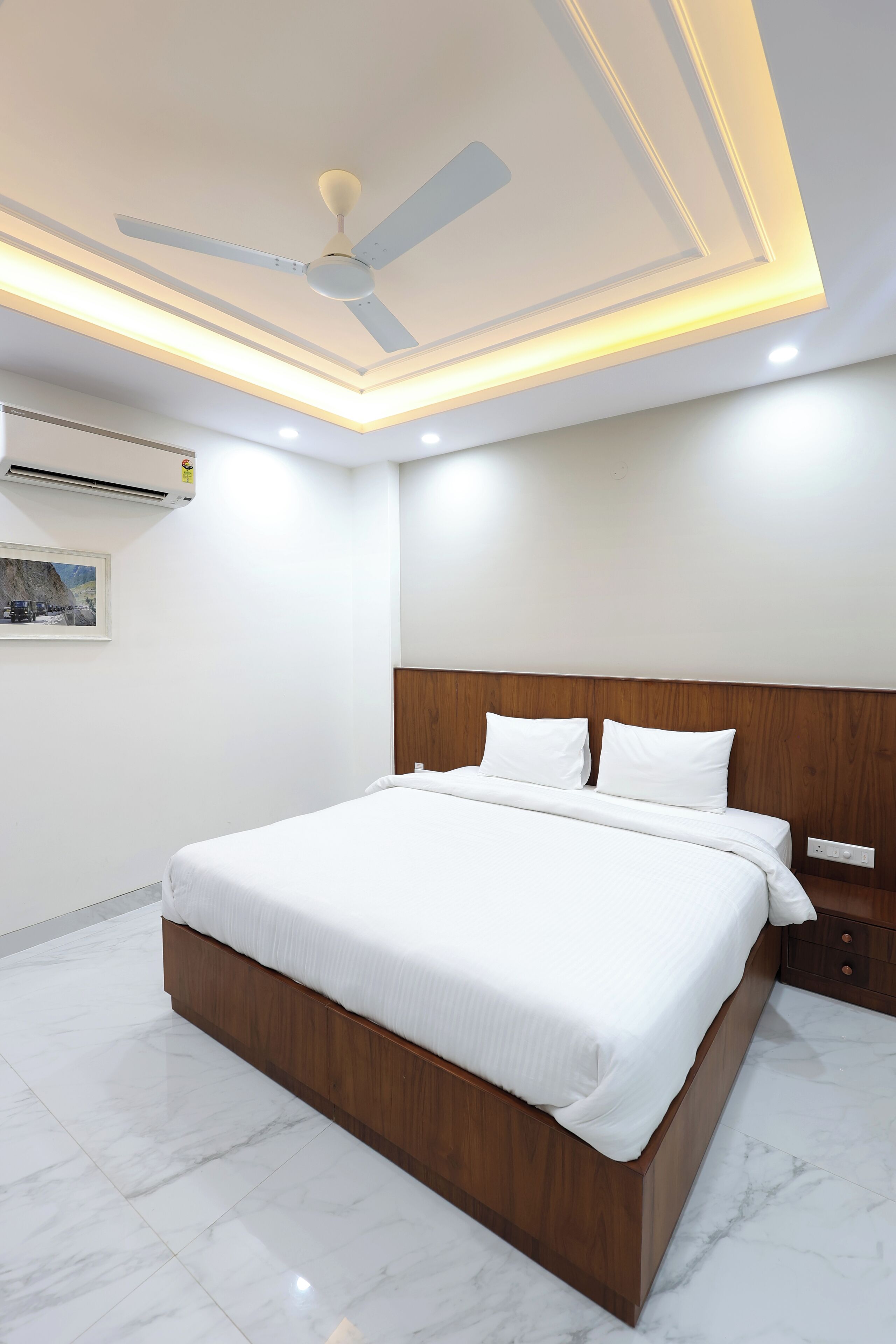 A modern bedroom with a wooden headboard, a large bed, a ceiling fan, recessed lighting, and a wall-mounted air conditioner.