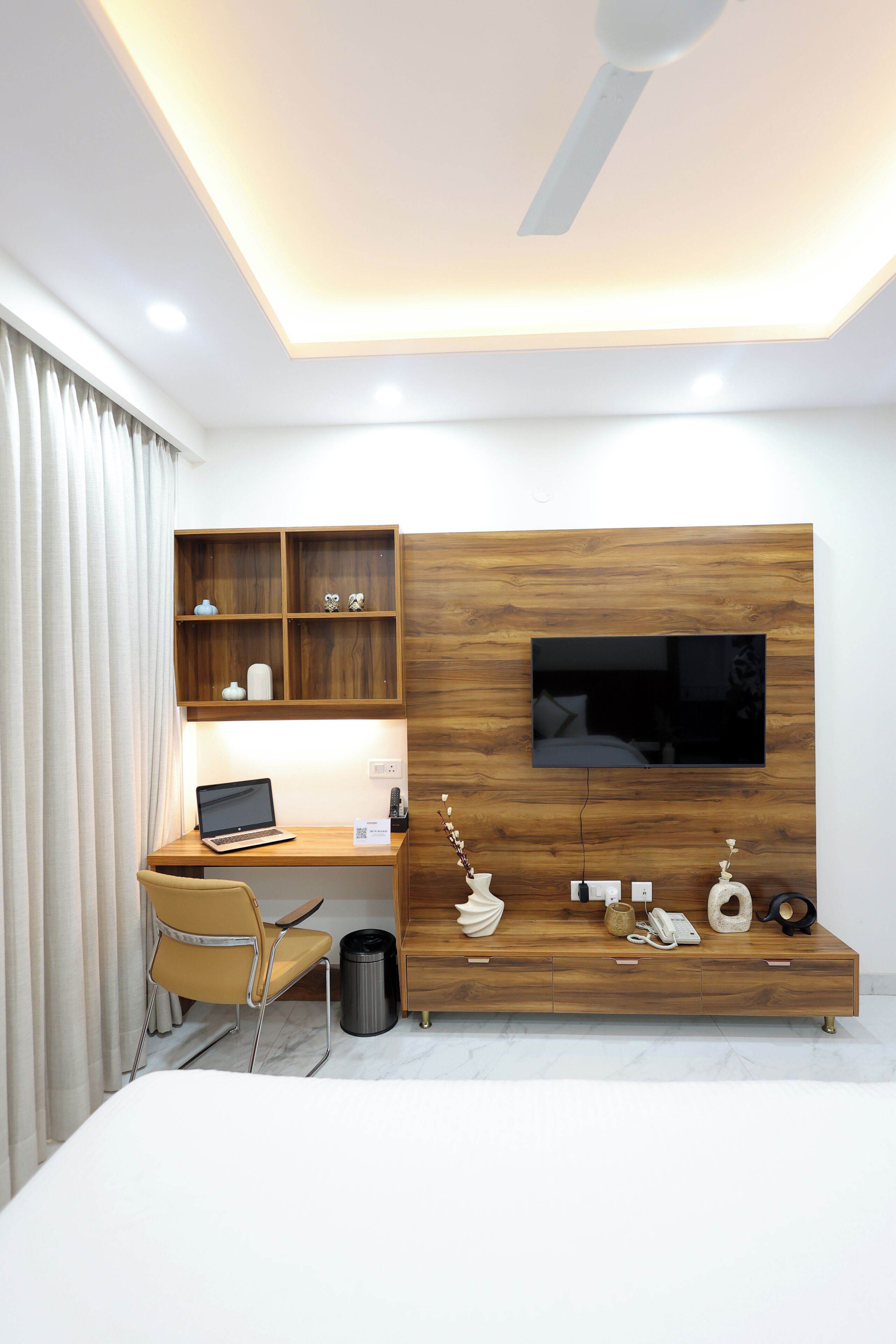 A modern bedroom with a wooden desk, a TV mounted on the wall, a chair, and a bedside table.