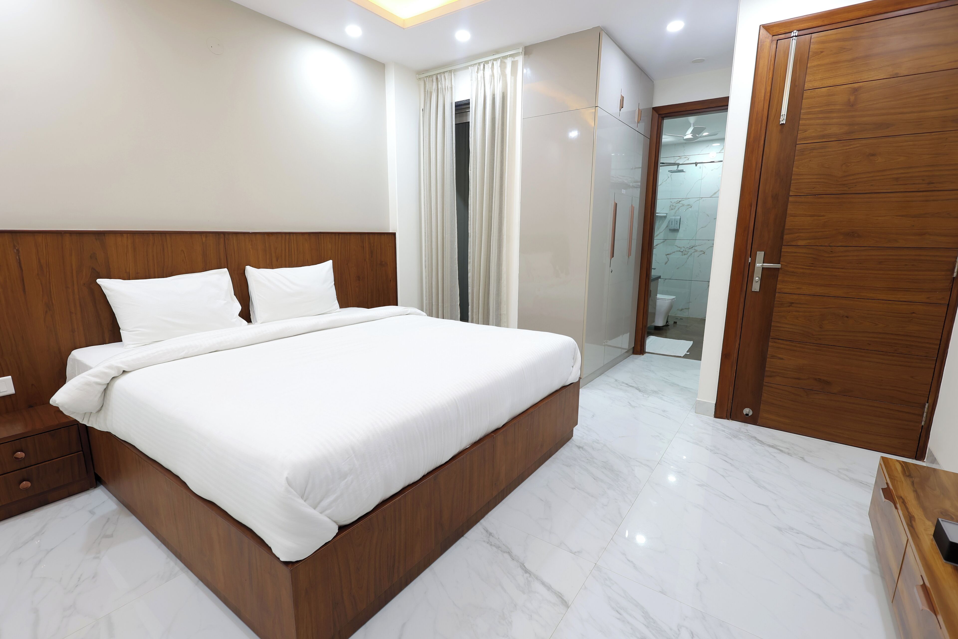 A modern hotel room with a large bed, wooden headboard, white linens, a wooden wardrobe, and a bathroom visible through a glass door.