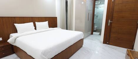 A modern hotel room with a large bed, wooden headboard, white linens, a wooden wardrobe, and a bathroom visible through a glass door.