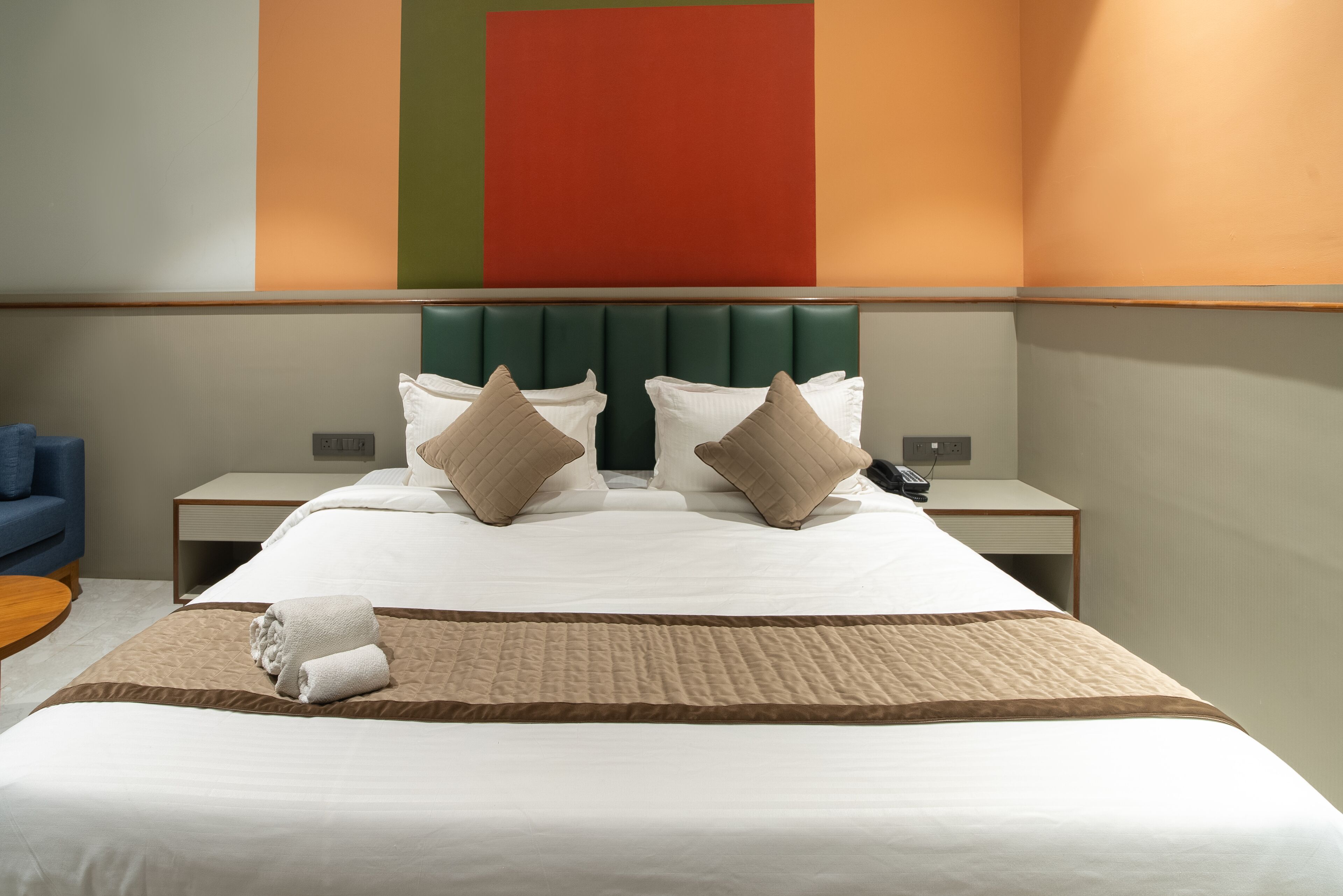 Una habitación de hotel con una cama grande, dos mesitas de noche, un sillón azul y una pared con un diseño geométrico.