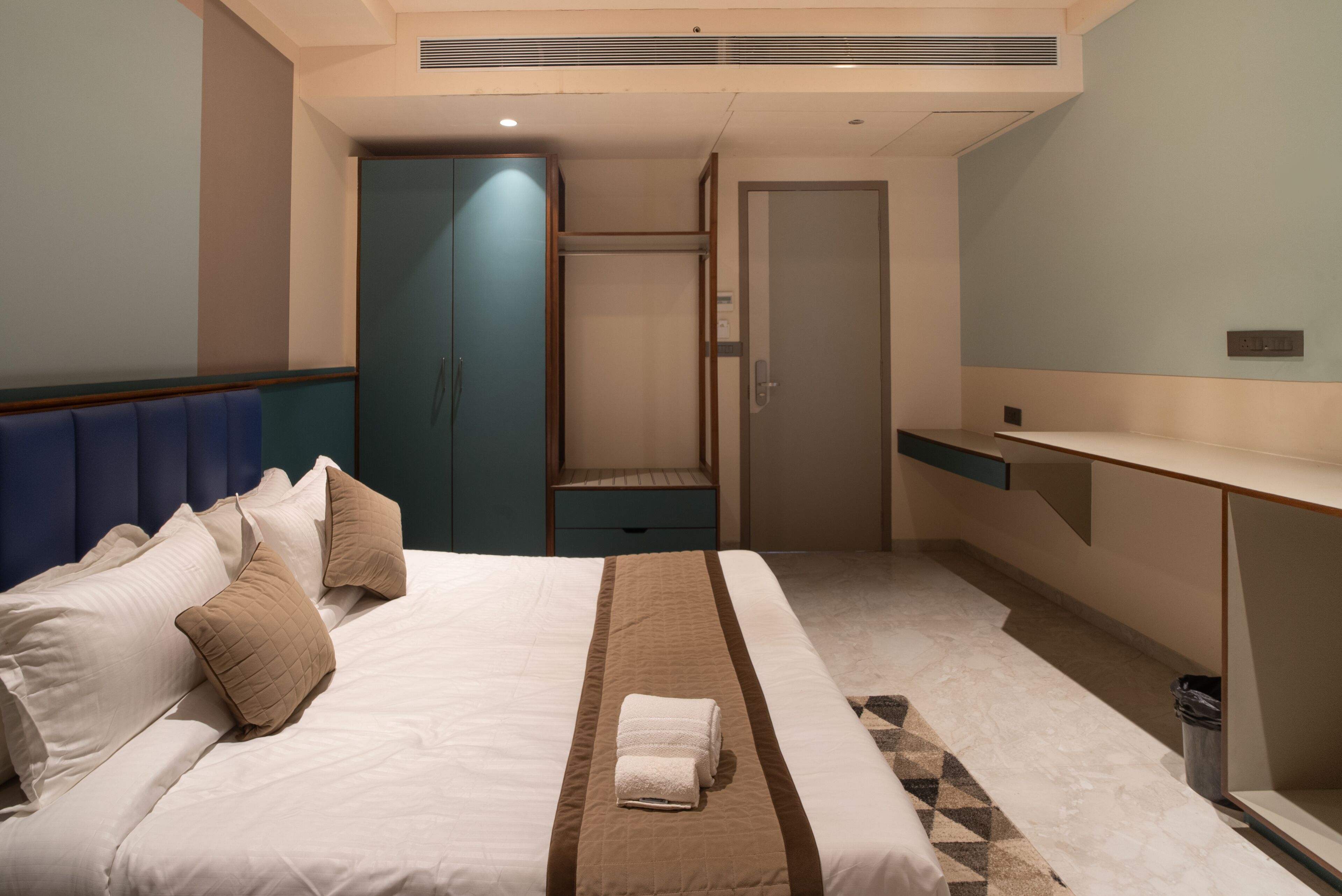 A hotel room with a bed, a wardrobe, a desk, and a trash bin.