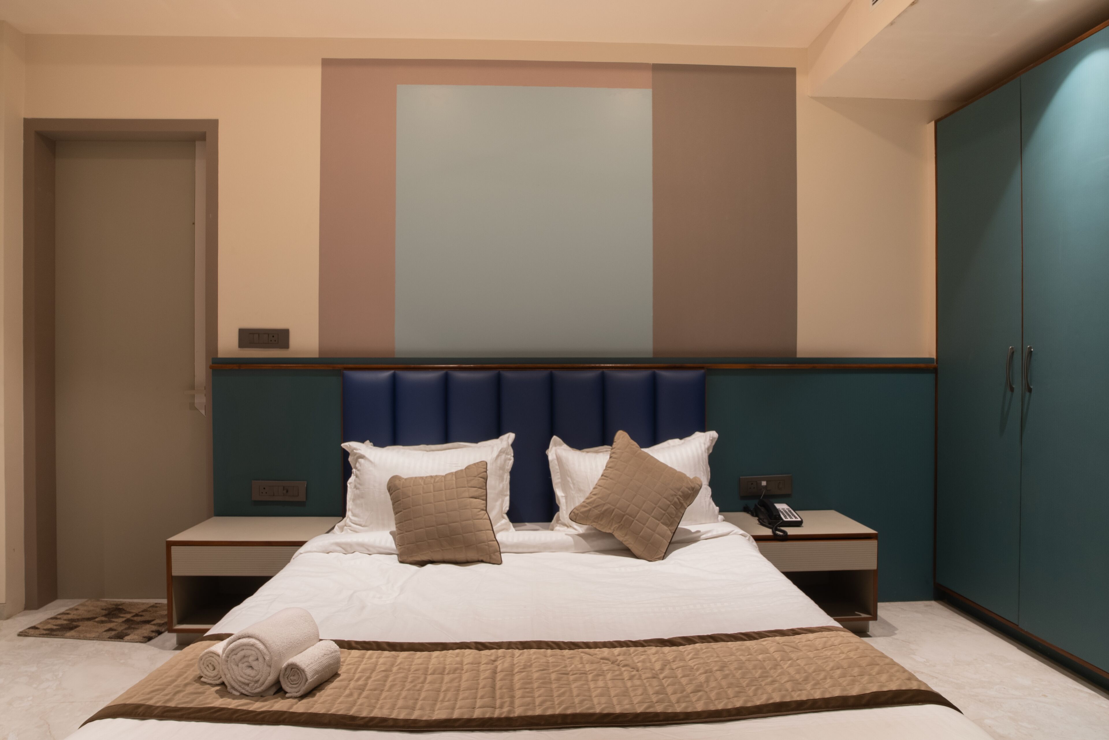 Una habitación de hotel moderna con una cama grande, cabecera con panel azul y una mesita de noche con teléfono, además de un armario.