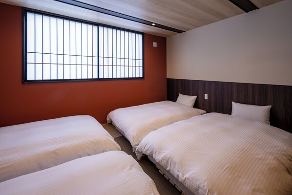 A room with three single beds, a large window with vertical blinds, and a wooden headboard.